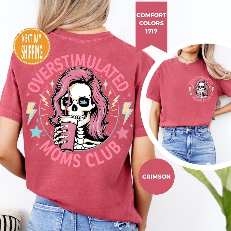 May include: Crimson-colored t-shirt featuring a graphic of a skull with pink hair, holding a cup, and the words "OVERSTIMULATED MOMS CLUB." The design includes lightning bolts and stars. The shirt is a comfort color.