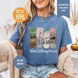 May include: Blue jean-colored t-shirt featuring a graphic of Walt Disney World landmarks. The design includes images of a castle, trees, and other architectural elements. The words "Walt Disney World" are printed in white script below the graphic. The shirt has a crew neck and short sleeves.