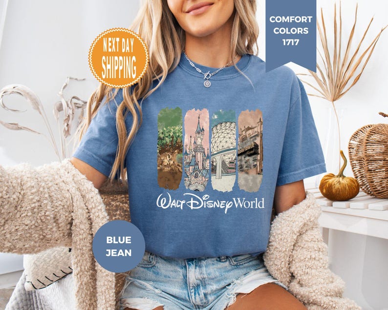 May include: Blue jean-colored t-shirt featuring a graphic of Walt Disney World landmarks. The design includes images of a castle, trees, and other architectural elements. The words "Walt Disney World" are printed in white script below the graphic. The shirt has a crew neck and short sleeves.