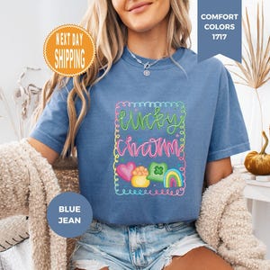 May include: Blue jean colored t-shirt with the words "Lucky Charm" in a colorful, whimsical design. The design includes a rainbow, hearts, and a pot of gold. The shirt is a Comfort Colors 1717 style.