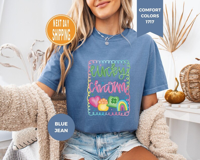 May include: Blue jean colored t-shirt with the words "Lucky Charm" in a colorful, whimsical design. The design includes a rainbow, hearts, and a pot of gold. The shirt is a Comfort Colors 1717 style.