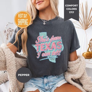 May include: A pepper-colored short-sleeve t-shirt featuring a distressed light blue map of Texas with pink and white text that reads She's from TEXAS I can tell. The shirt has a crew neckline and a relaxed fit, showcasing a vintage-inspired graphic.