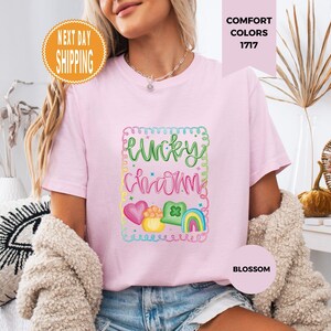 May include: A light pink t-shirt with the words "Lucky Charm" in green and pink script, surrounded by a colorful frame with hearts, a pot of gold, a shamrock, and a rainbow. The shirt is labeled "Blossom" and "Comfort Colors 1717".