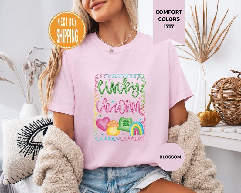 May include: A light pink t-shirt with the words "Lucky Charm" in green and pink script, surrounded by a colorful frame with hearts, a pot of gold, a shamrock, and a rainbow. The shirt is labeled "Blossom" and "Comfort Colors 1717".