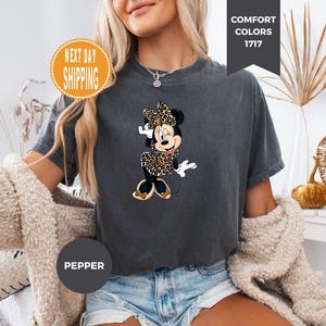 May include: Dark gray t-shirt featuring a Minnie Mouse graphic in a leopard print design. The shirt has short sleeves and a crew neck. The text "Comfort Colors 1717" is on the top right. The text "Pepper" is on the bottom left. The text "Next Day Shipping" is on the top left.