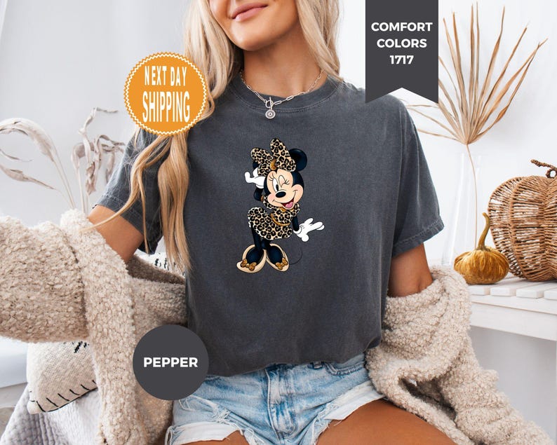 May include: Dark gray t-shirt featuring a Minnie Mouse graphic in a leopard print design. The shirt has short sleeves and a crew neck. The text "Comfort Colors 1717" is on the top right. The text "Pepper" is on the bottom left. The text "Next Day Shipping" is on the top left.