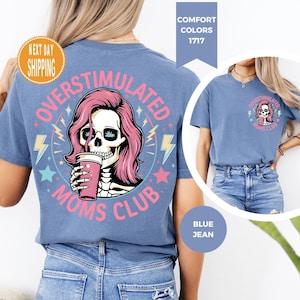 May include: Blue jean-colored t-shirt with a graphic on the front and back. The graphic features a skull with pink hair holding a cup, surrounded by text that reads "OVERSTIMULATED MOMS CLUB". The shirt has a crew neck and short sleeves.