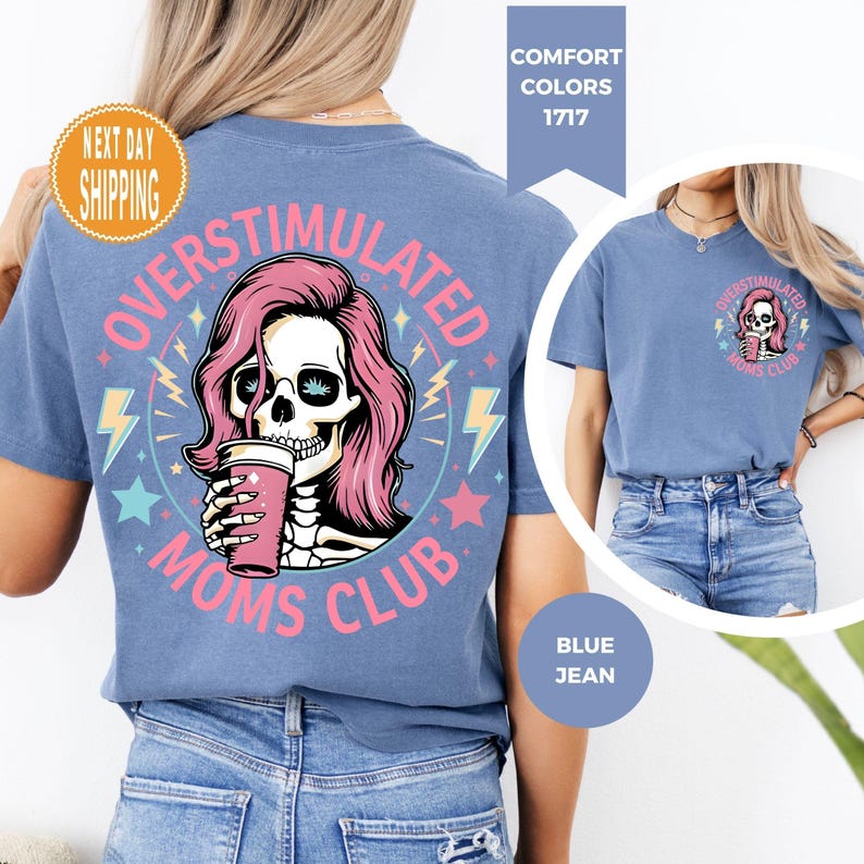 May include: Blue jean-colored t-shirt with a graphic on the front and back. The graphic features a skull with pink hair holding a cup, surrounded by text that reads "OVERSTIMULATED MOMS CLUB". The shirt has a crew neck and short sleeves.