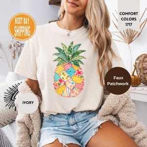 May include: An ivory t-shirt featuring a central graphic of a pineapple designed with a faux patchwork pattern. The pineapple displays various colourful segments including yellow, pink, and blue floral and geometric prints with green leaves.