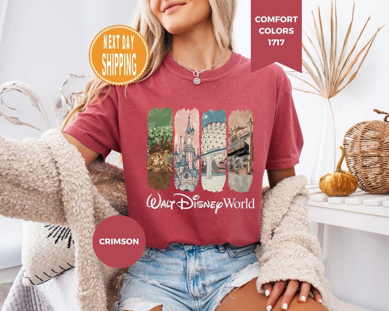 May include: Crimson-colored t-shirt featuring a graphic of Walt Disney World landmarks. The shirt has a crew neck and short sleeves. The words "Walt Disney World" are printed in white script below the graphic. The shirt is made of a soft material.