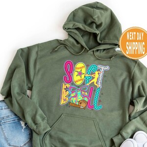 May include: Olive green hooded sweatshirt with a colorful "Softball" graphic. The graphic includes a yellow softball with a star, a batter, and a baseball bat. The words are in pink, blue, and green.