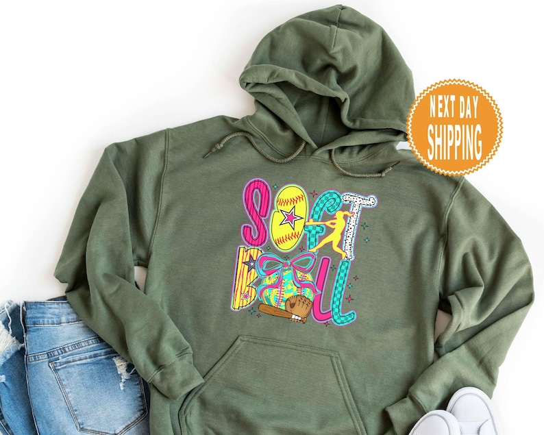 May include: Olive green hooded sweatshirt with a colorful "Softball" graphic. The graphic includes a yellow softball with a star, a batter, and a baseball bat. The words are in pink, blue, and green.