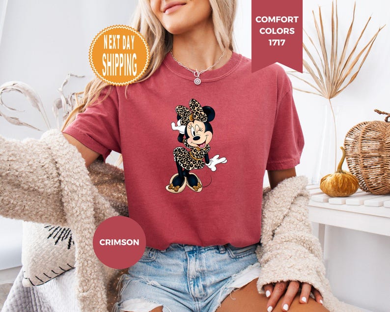May include: Crimson-colored t-shirt featuring a Minnie Mouse graphic in a leopard print dress and bow. The shirt has short sleeves and is paired with denim shorts. The words "COMFORT COLORS 1717" are displayed on a banner.