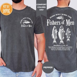 May include: Dark grey t-shirt with white text and graphics. The front features a fishing lure design and the words "Jesus Will Make You Fishers of Men." The back displays three fish and the same text, along with a Bible verse.