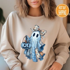 Op de afbeelding: Beige sweatshirt met een aquarel illustratie van een vriendelijke geest met een blauwe pompoenemmer. De geest heeft een bloemenstrik en patches. Inclusief een "NEXT DAY SHIPPING" sticker.