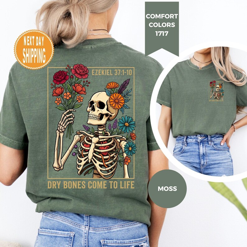 May include: A moss green t-shirt featuring a skeleton holding a bouquet of flowers, with the text "DRY BONES COME TO LIFE" and "EZEKIEL 37:1-10". The shirt also has a small graphic on the upper back. The shirt is part of the Comfort Colors 1717 collection.