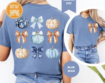 Könnte beinhalten: Ein blaues T-Shirt mit einer Grafik auf der Rückseite, die neun abwechselnde Kürbisse und Schleifen in Blau, Weiß und Orange mit Blumen- und Vichy-Mustern zeigt. Auf der linken Brustseite befindet sich eine einzelne orange-weiße Schleife.