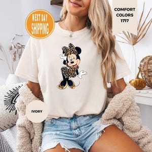 May include: Ivory-colored t-shirt featuring a Minnie Mouse graphic in a leopard print design. The shirt has short sleeves and a crew neck. The words "COMFORT COLORS 1717" are printed on the shirt. The shirt is worn by a person.