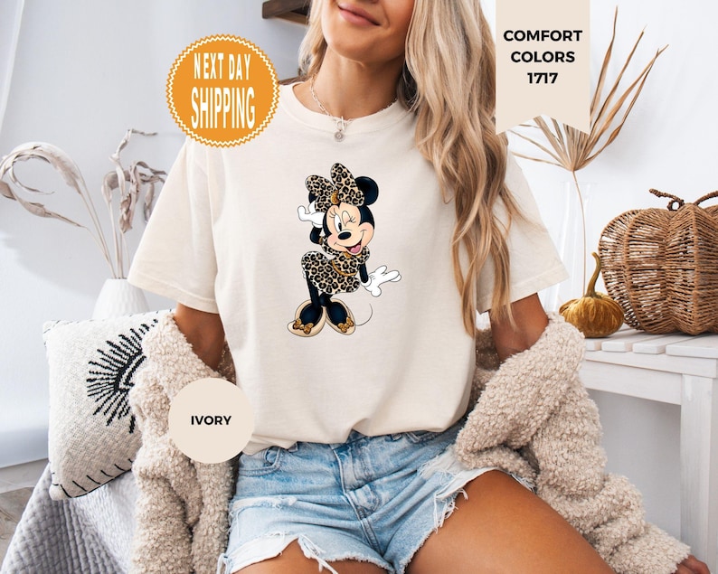 May include: Ivory-colored t-shirt featuring a Minnie Mouse graphic in a leopard print design. The shirt has short sleeves and a crew neck. The words "COMFORT COLORS 1717" are printed on the shirt. The shirt is worn by a person.