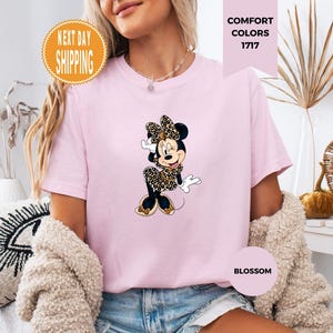 May include: Light pink t-shirt featuring a Minnie Mouse graphic in a leopard print bow and dress. The shirt has the text "Comfort Colors 1717" and "Blossom". An orange circle with the text "Next Day Shipping" is also visible.