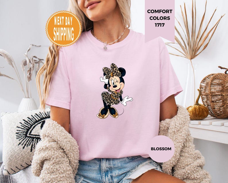 May include: Light pink t-shirt featuring a Minnie Mouse graphic in a leopard print bow and dress. The shirt has the text "Comfort Colors 1717" and "Blossom". An orange circle with the text "Next Day Shipping" is also visible.