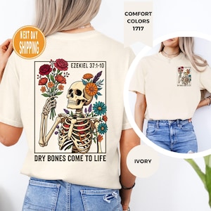 May include: Ivory-colored t-shirt featuring a skeletal figure holding a bouquet of flowers. The design includes the text "EZEKIEL 37:1-10" and "DRY BONES COME TO LIFE" in a black rectangular frame. The shirt also has a small design on the front.