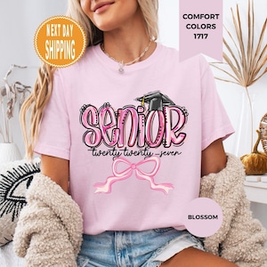May include: A light pink t-shirt featuring a graphic with the word SENIOR in bubble letters, a graduation cap, the text twenty twenty-seven, and a pink ribbon bow. The shirt is labeled Comfort Colors 1717 and the color is identified as Blossom.