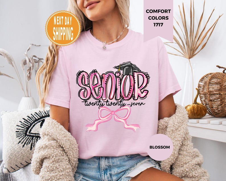 May include: A light pink t-shirt featuring a graphic with the word SENIOR in bubble letters, a graduation cap, the text twenty twenty-seven, and a pink ribbon bow. The shirt is labeled Comfort Colors 1717 and the color is identified as Blossom.