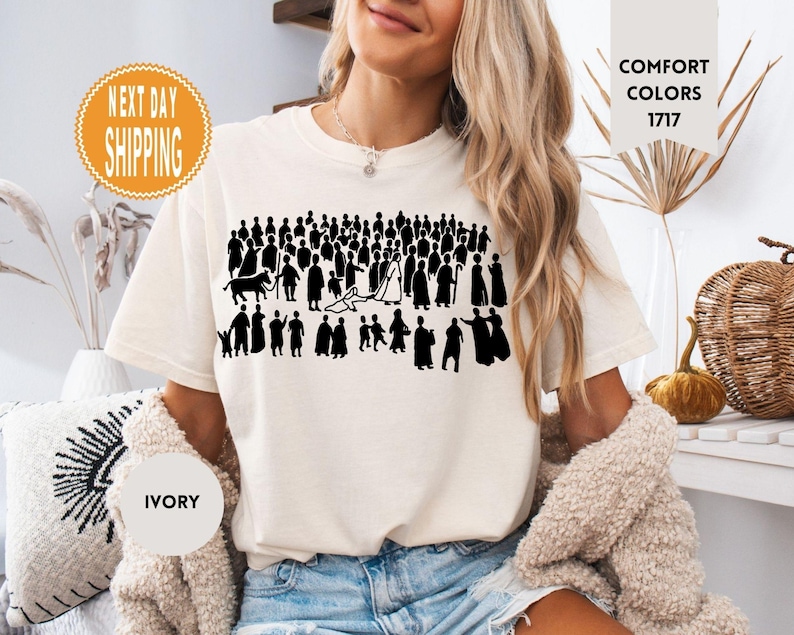 May include: Ivory-colored t-shirt featuring a black silhouette of a crowd of people. The shirt has the words "Comfort Colors 1717" printed on it. The shirt also has the words "Next Day Shipping" printed on it.