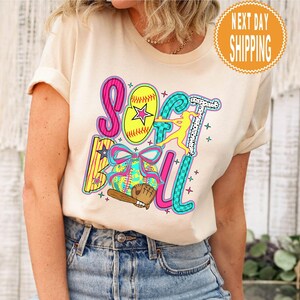 May include: A cream-colored t-shirt featuring the word "SOFTBALL" in colorful, playful lettering. The design includes a softball, a baseball glove, a bat, and a silhouetted softball player. A round orange sticker with the words "NEXT DAY SHIPPING" is visible.
