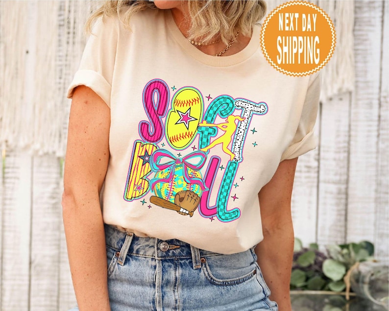 May include: A cream-colored t-shirt featuring the word "SOFTBALL" in colorful, playful lettering. The design includes a softball, a baseball glove, a bat, and a silhouetted softball player. A round orange sticker with the words "NEXT DAY SHIPPING" is visible.