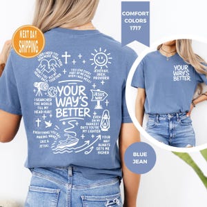 May include: A blue jean-coloured t-shirt with the text "YOUR WAY'S BETTER" in white. The back of the shirt features a graphic design with religious phrases and symbols, including a sun, cross, and boat. Part of the "Comfort Colors" collection.