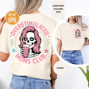 May include: Ivory-colored t-shirt featuring a graphic of a skull with pink hair holding a pink cup, surrounded by the words "OVERSTIMULATED MOMS CLUB." The design includes stars and lightning bolts. The shirt is a comfort color.