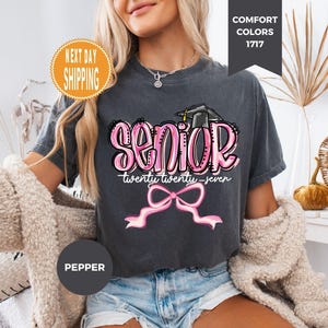 May include: A charcoal grey t-shirt featuring the word SENIOR in large pink bubble letters with a black graduation cap on top. Below, the text reads twenty twenty-seven in white script, followed by a large pink ribbon bow graphic.