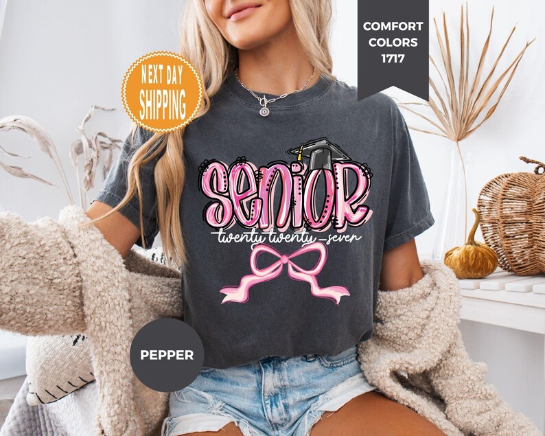 May include: A charcoal grey t-shirt featuring the word SENIOR in large pink bubble letters with a black graduation cap on top. Below, the text reads twenty twenty-seven in white script, followed by a large pink ribbon bow graphic.