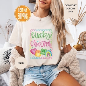 May include: Ivory-colored t-shirt with a colorful "Lucky Charm" graphic. The design includes a heart, pot of gold, shamrock, and rainbow. The shirt is part of the Comfort Colors collection.
