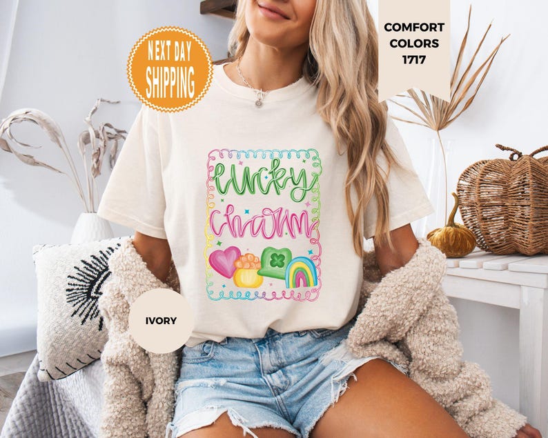 May include: Ivory-colored t-shirt with a colorful "Lucky Charm" graphic. The design includes a heart, pot of gold, shamrock, and rainbow. The shirt is part of the Comfort Colors collection.