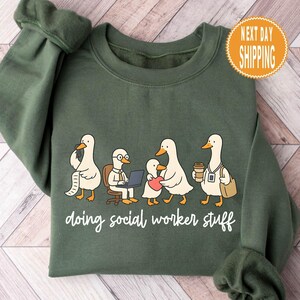 Könnte beinhalten: Ein grünes Sweatshirt mit einer Cartoon-Illustration von fünf Enten. Die Enten werden in verschiedenen Szenarien dargestellt, darunter die Benutzung eines Laptops und das Tragen einer Kaffeetasse. Der Text "doing social worker stuff" steht in weißer Schrift unten.
