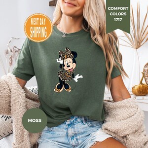 May include: Moss green t-shirt featuring a Minnie Mouse graphic in a leopard print dress and bow. The shirt has the words "Comfort Colors 1717" and "Moss" printed on it. The shirt is a relaxed fit.