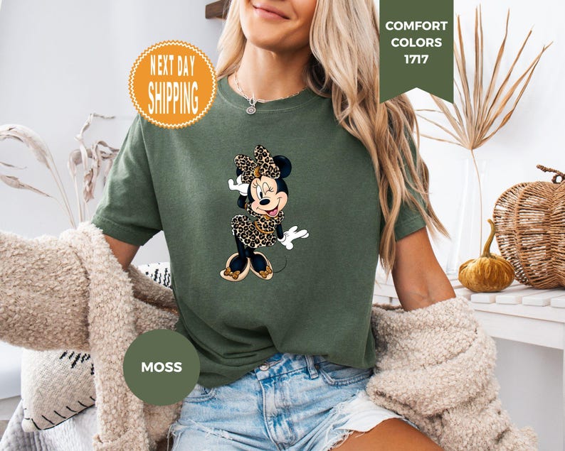 May include: Moss green t-shirt featuring a Minnie Mouse graphic in a leopard print dress and bow. The shirt has the words "Comfort Colors 1717" and "Moss" printed on it. The shirt is a relaxed fit.