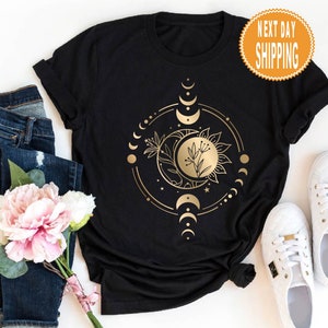 Op de afbeelding: Zwart T-shirt met een gouden ontwerp van een zon en maan met een bloemelement in het midden. Het ontwerp is omgeven door een cirkel van kleinere manen en sterren. De tekst "Next Day Shipping" staat op een gouden sticker in de rechterbovenhoek van de afbeelding.