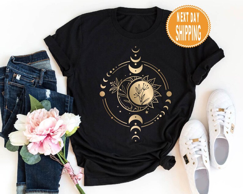 Op de afbeelding: Zwart T-shirt met een gouden ontwerp van een zon en maan met een bloemelement in het midden. Het ontwerp is omgeven door een cirkel van kleinere manen en sterren. De tekst "Next Day Shipping" staat op een gouden sticker in de rechterbovenhoek van de afbeelding.
