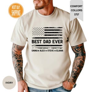 May include: An ivory short-sleeve t-shirt featuring a black distressed American flag graphic. The text BEST DAD EVER is printed in the center, followed by the names EMMA, ALEX, STEVE, and ELIANA separated by stars in a horizontal line.