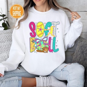 May include: White sweatshirt with a colorful "Softball" graphic. The design includes a yellow softball with a star, a batter, a glove, and the word "Softball" in pink, teal, and yellow. The sweatshirt is worn by a person in blue jeans.
