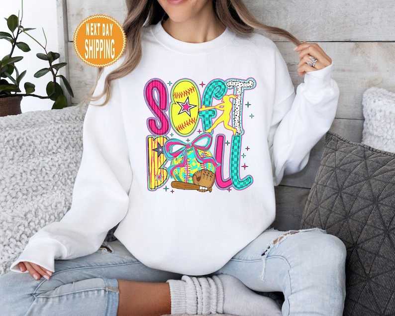 May include: White sweatshirt with a colorful "Softball" graphic. The design includes a yellow softball with a star, a batter, a glove, and the word "Softball" in pink, teal, and yellow. The sweatshirt is worn by a person in blue jeans.