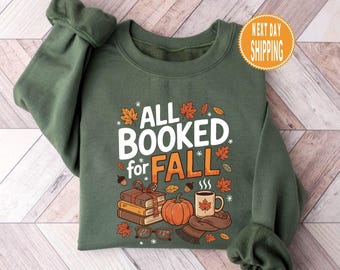 Op de afbeelding: Een groene sweatshirt met de tekst "ALL BOOKED for FALL" in wit en oranje. Het ontwerp bevat illustraties van boeken, een pompoen, een koffiemok en herfstbladeren. De sweatshirt heeft lange mouwen en een ronde hals.