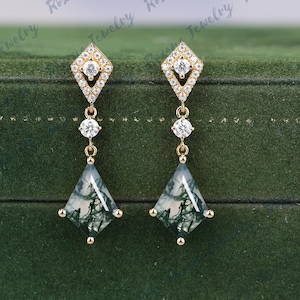 Op de afbeelding: Goudkleurige oorhangers met een ruitvormige bovenkant en een met diamanten bezet ontwerp. De oorbellen hebben een trio van ronde, heldere stenen en een groen-witte driehoekige steen aan de onderkant. De oorbellen worden gepresenteerd op groen fluweel.