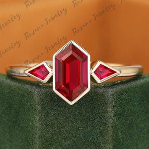 May include: A gold ring with a large, rectangular, red gemstone in the center, flanked by two smaller, triangular red gemstones. The ring has a delicate, elegant design. The text "Roxana Jewelry" is visible in the background.