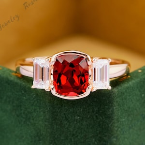 May include: A rose gold engagement ring with a large, square-shaped red gemstone surrounded by two smaller, rectangular-shaped white gemstones.