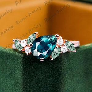 Può includere: Un anello in oro rosa con una gemma a forma di pera di colore teal come elemento centrale. L'anello è impreziosito da piccole gemme rotonde e trasparenti e da dettagli verdi a forma di foglia. Presentato su uno sfondo di velluto verde scuro.